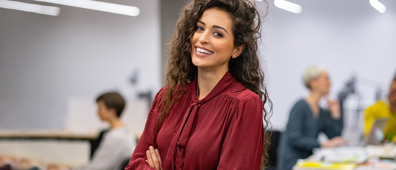 Successful businesswoman standing in creative office and looking at camera. Young latin woman entrepreneur in a coworking space smiling. Portrait of beautiful business woman standing in front of business team at modern agency with copy space.